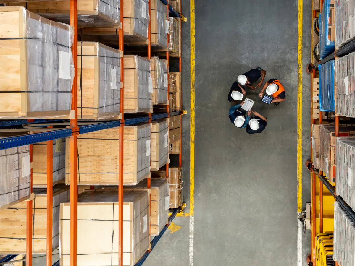 Overhead view of four warehouse workers in hard hats and safety vests gathered around a tablet in an aisle between pallet racks
