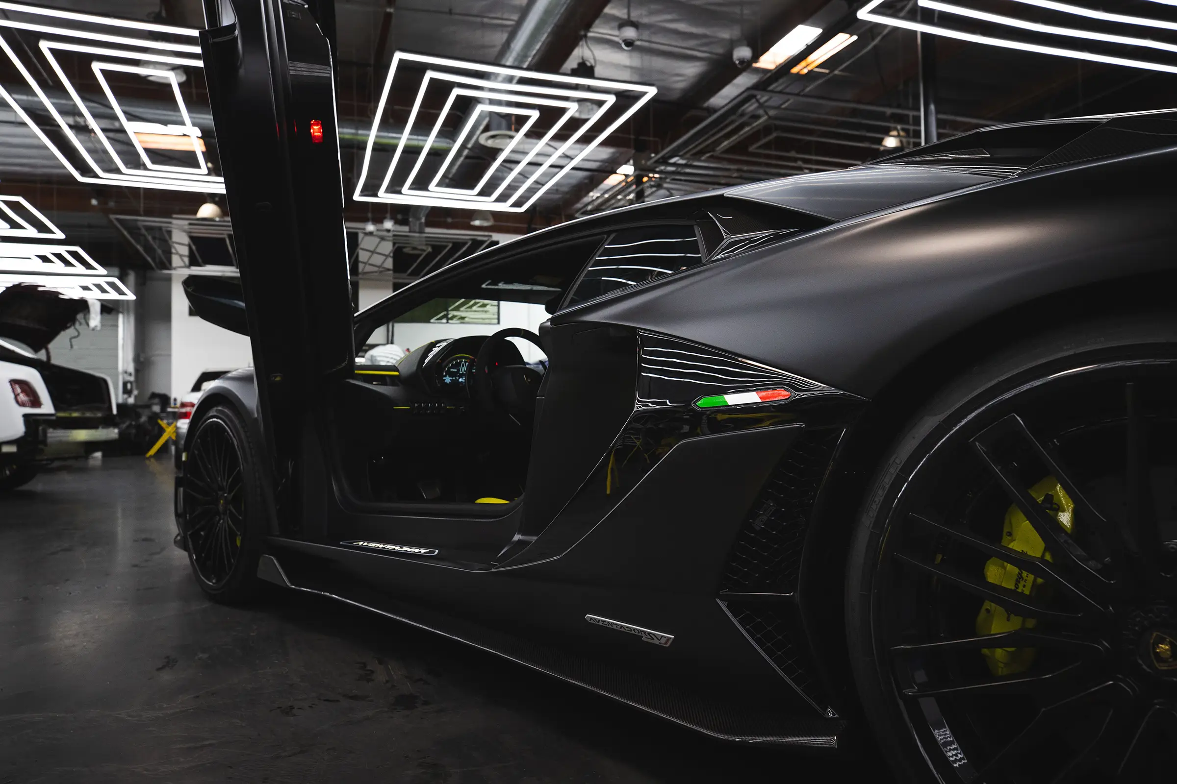 Matte black Lamborghini sports car with butterfly door open, showing part of the interior; yellow brake calipers and Italian flag emblem visible on the side in a garage with modern ceiling lights.