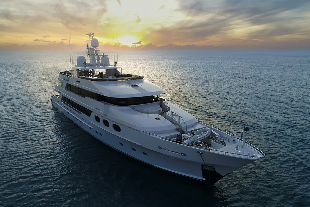 Large white luxury yacht floating on calm ocean water during a colorful sunset with clouds.
