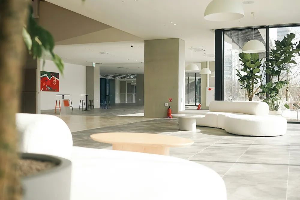 Bright modern indoor lounge area with curved white sofas, round coffee tables, large floor-to-ceiling windows letting in natural light, green potted plants, and minimalist ceiling lamps.