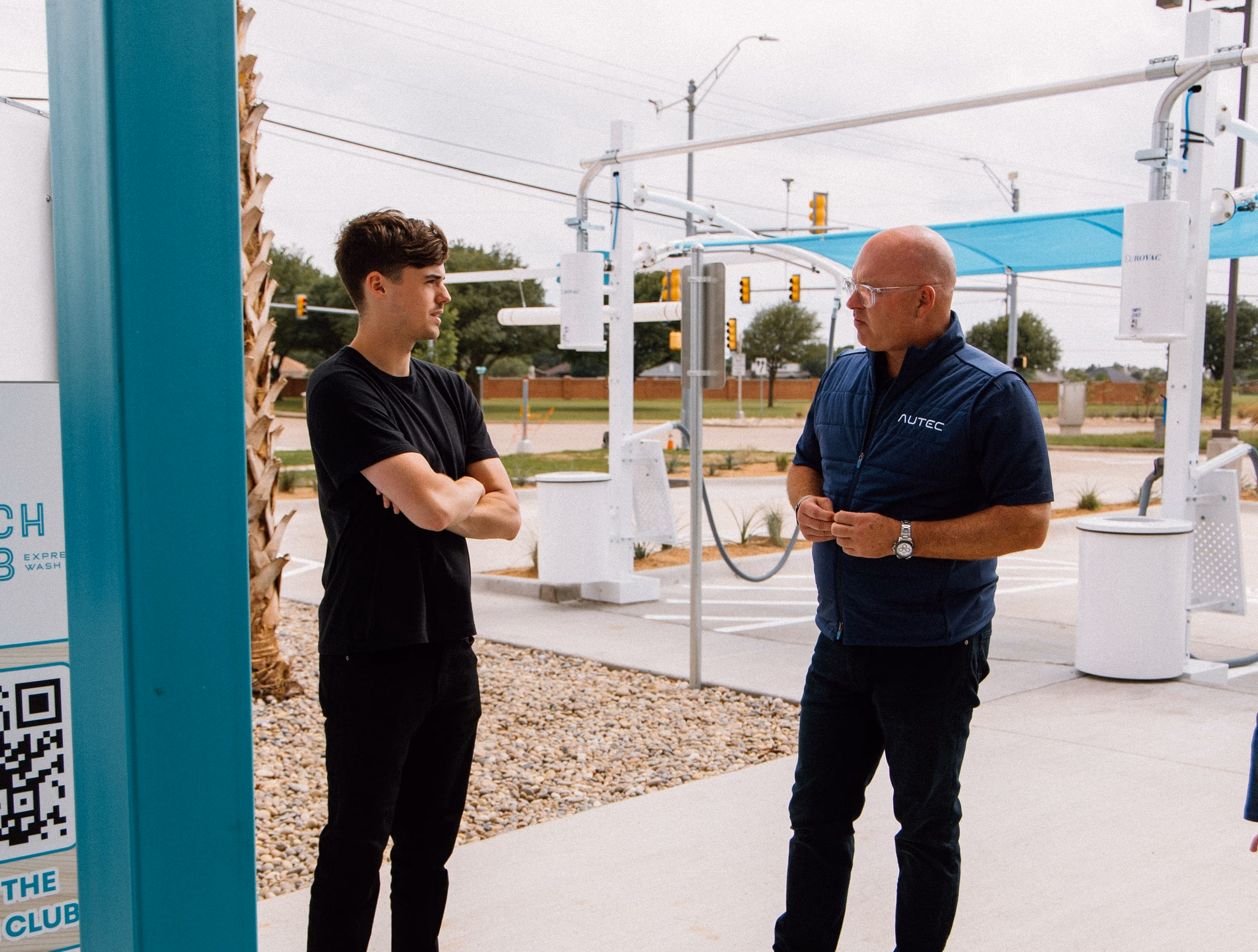 Two men standing outside near a car wash, one with arms crossed, the other in a vest labeled AUTEC.