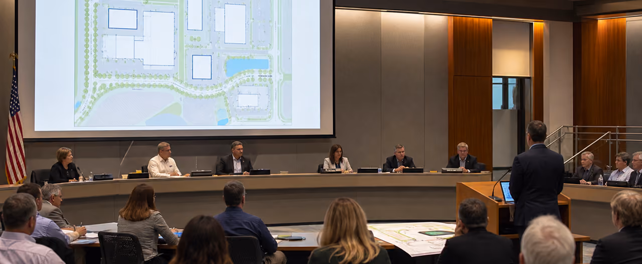 Man presenting urban planning maps to seated council members in a modern meeting room.