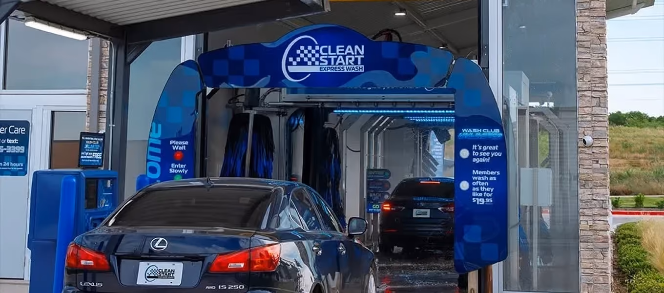 Rear view of a dark blue Lexus sedan positioned at the entrance of an automated car wash tunnel under a blue "Clean Start Express Wash" branded archway. A self-service payment kiosk is visible on the left, and another dark car is visible inside the wash tunnel ahead.