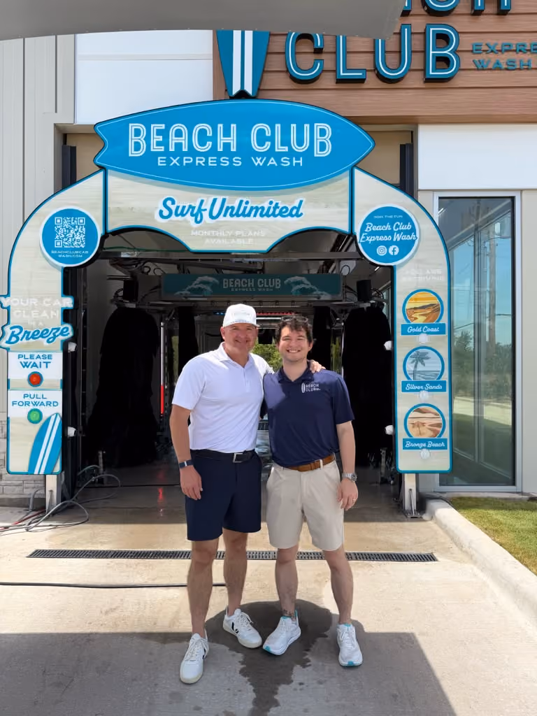 Two smiling men posing together outdoors in front of the entrance of a Beach Club Express Wash. The man on the left wears a white cap, white polo shirt, and navy shorts. The man on the right wears a navy polo shirt with a logo and beige shorts. Above them is a blue surfboard-shaped sign for "Beach Club Express Wash."