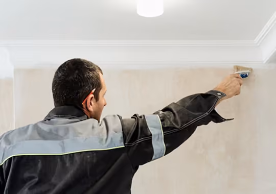 Man in dark work jacket painting a beige wall near the ceiling with a roller.