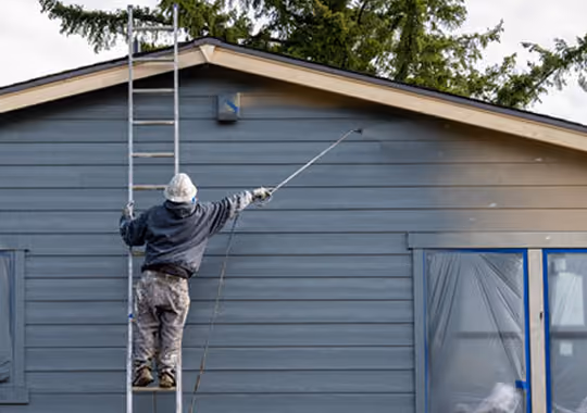 Person painting the exterior wall of a house gray while standing on a ladder.