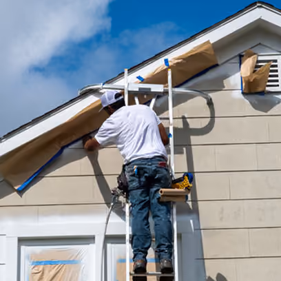 Man on ladder painting or repairing the exterior of a house under a blue sky.
