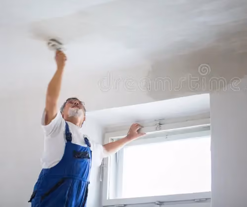 Man in blue overalls smoothing white ceiling plaster near a window.