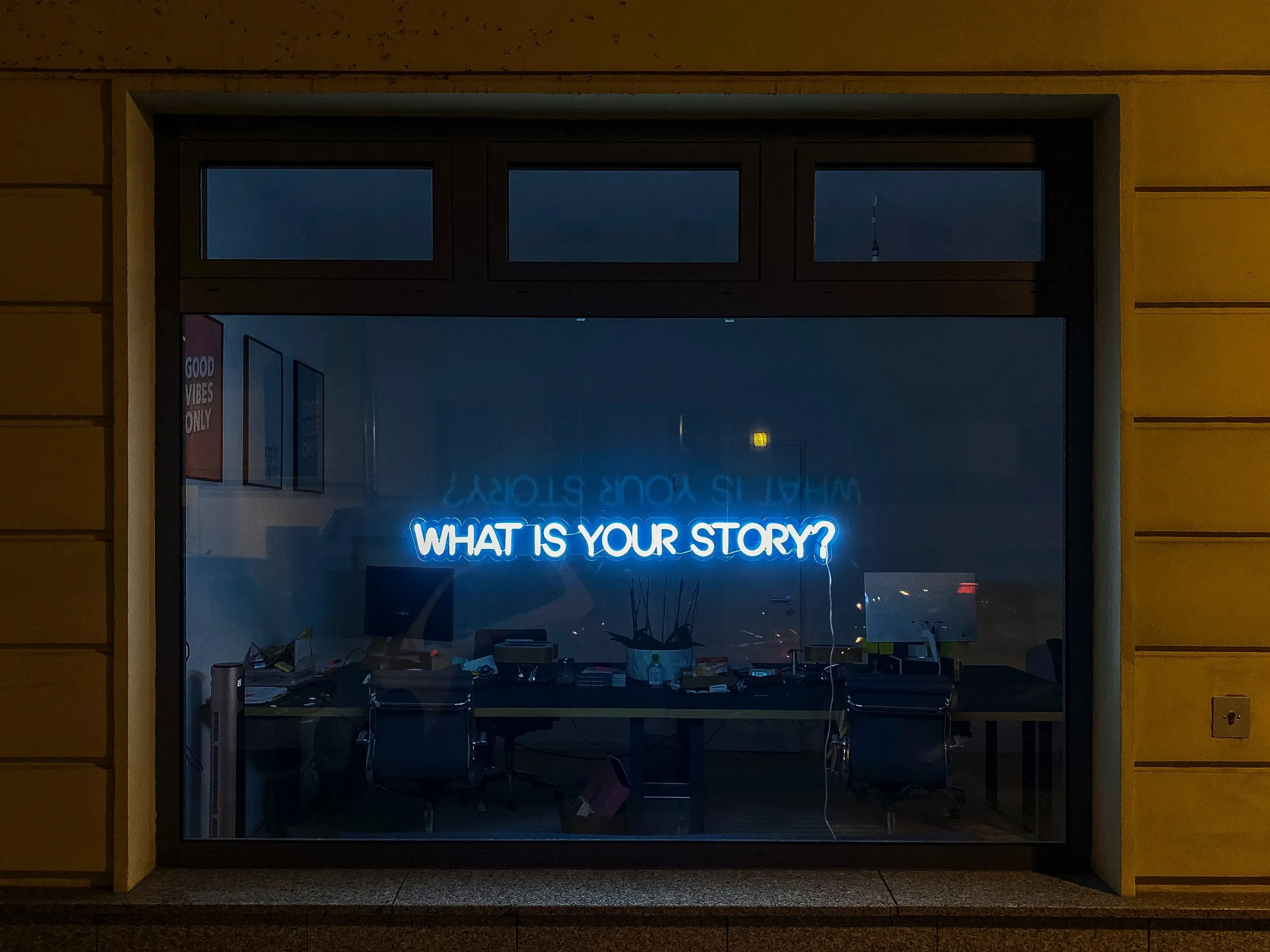 A neon sign on a window that says "What is your story?"