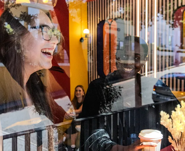 Two women smiling and laughing inside a cafe, one holding a coffee cup near a window