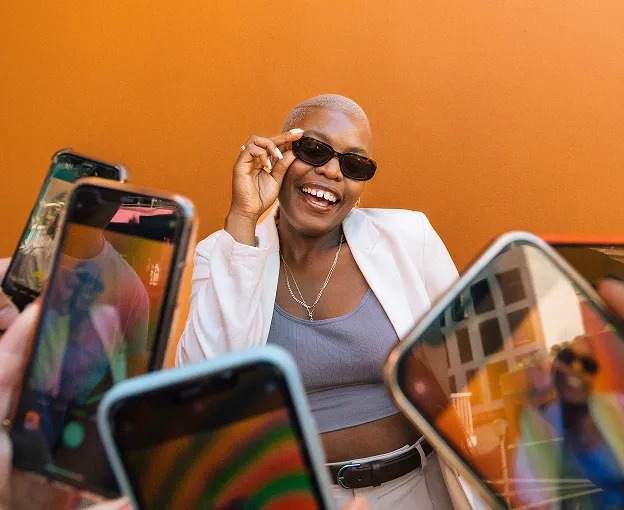 Smiling person wearing sunglasses and a white jacket posing against an orange wall while phones capture photos