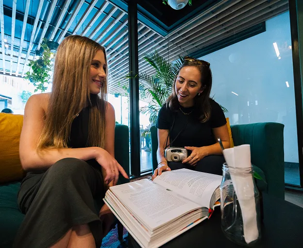 Two women sitting on couches looking at an open book in a modern indoor space with plants and glass walls