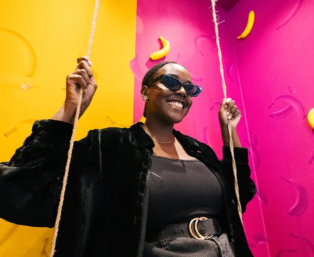 Smiling woman wearing sunglasses on a swing with yellow and pink walls featuring banana shapes