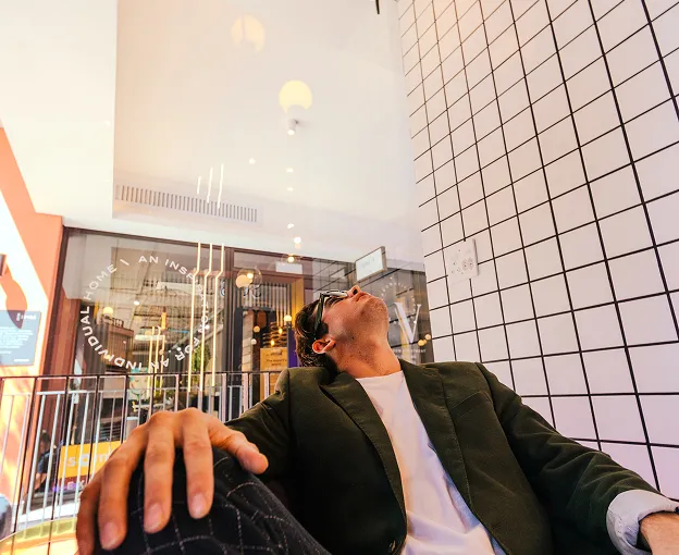 Man wearing sunglasses leaning back indoors against a white tiled wall with black grid lines