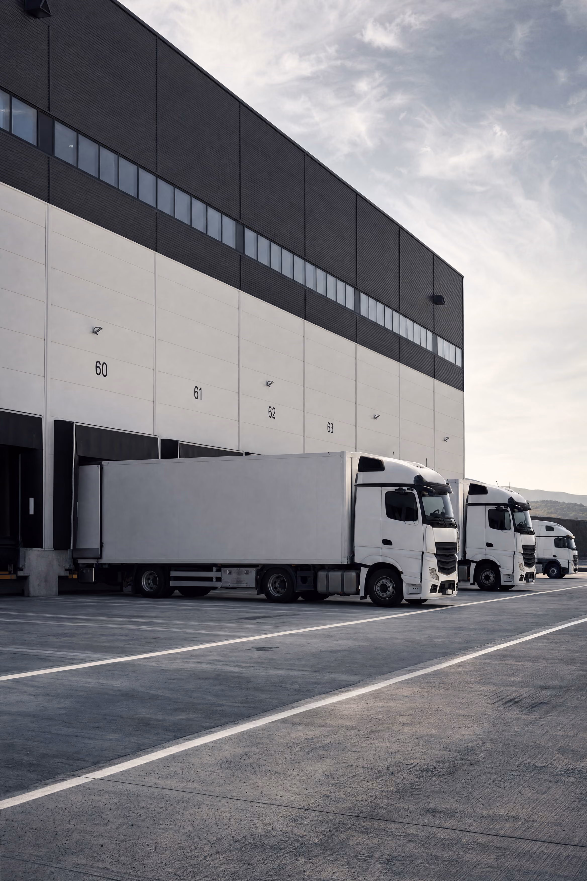 Three white semi-trailer trucks parked at loading docks numbered 60 to 63 at a warehouse.