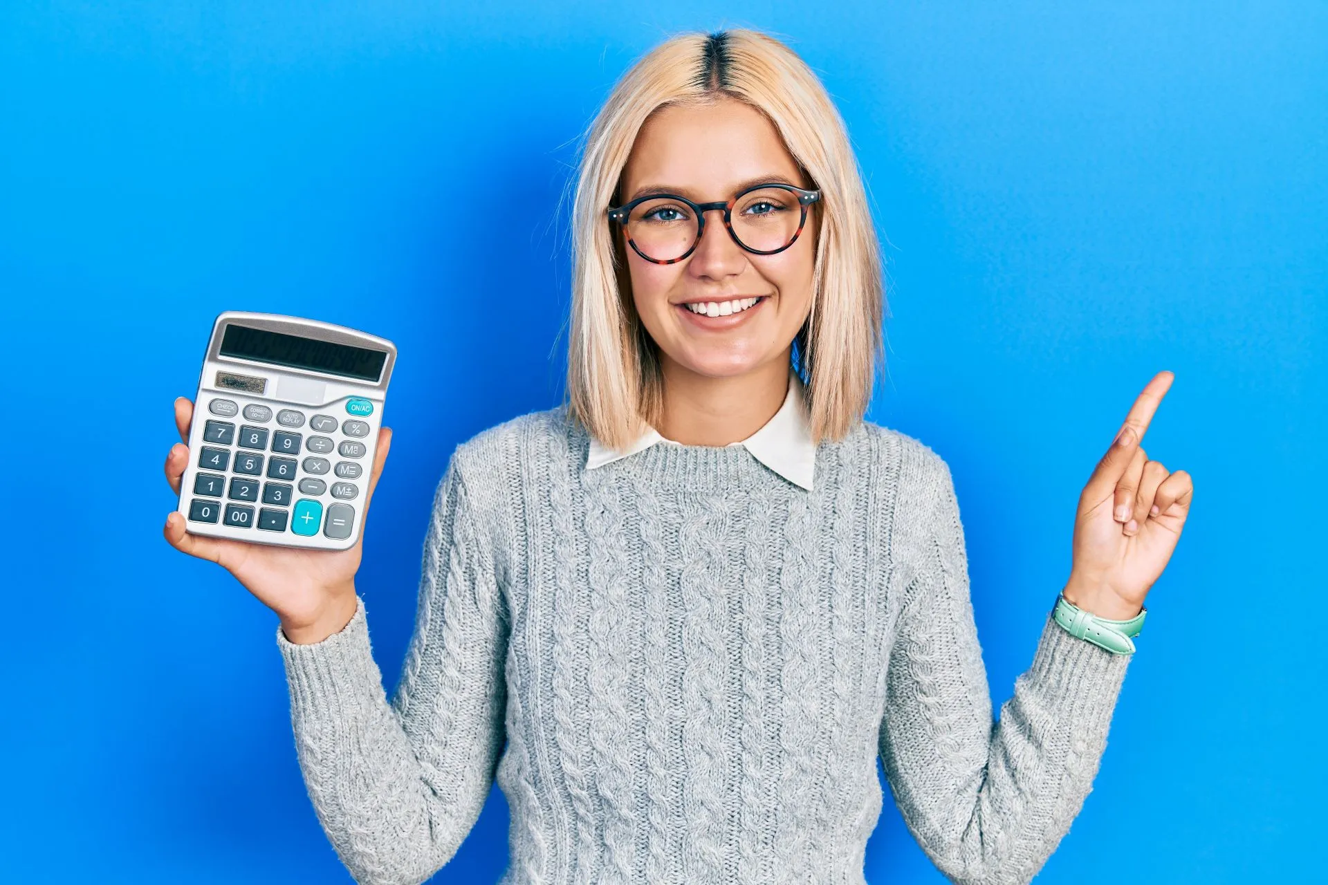 Smiling woman with blonde hair and glasses wearing a gray sweater holds a calculator in one hand and points up with the other against a blue background.