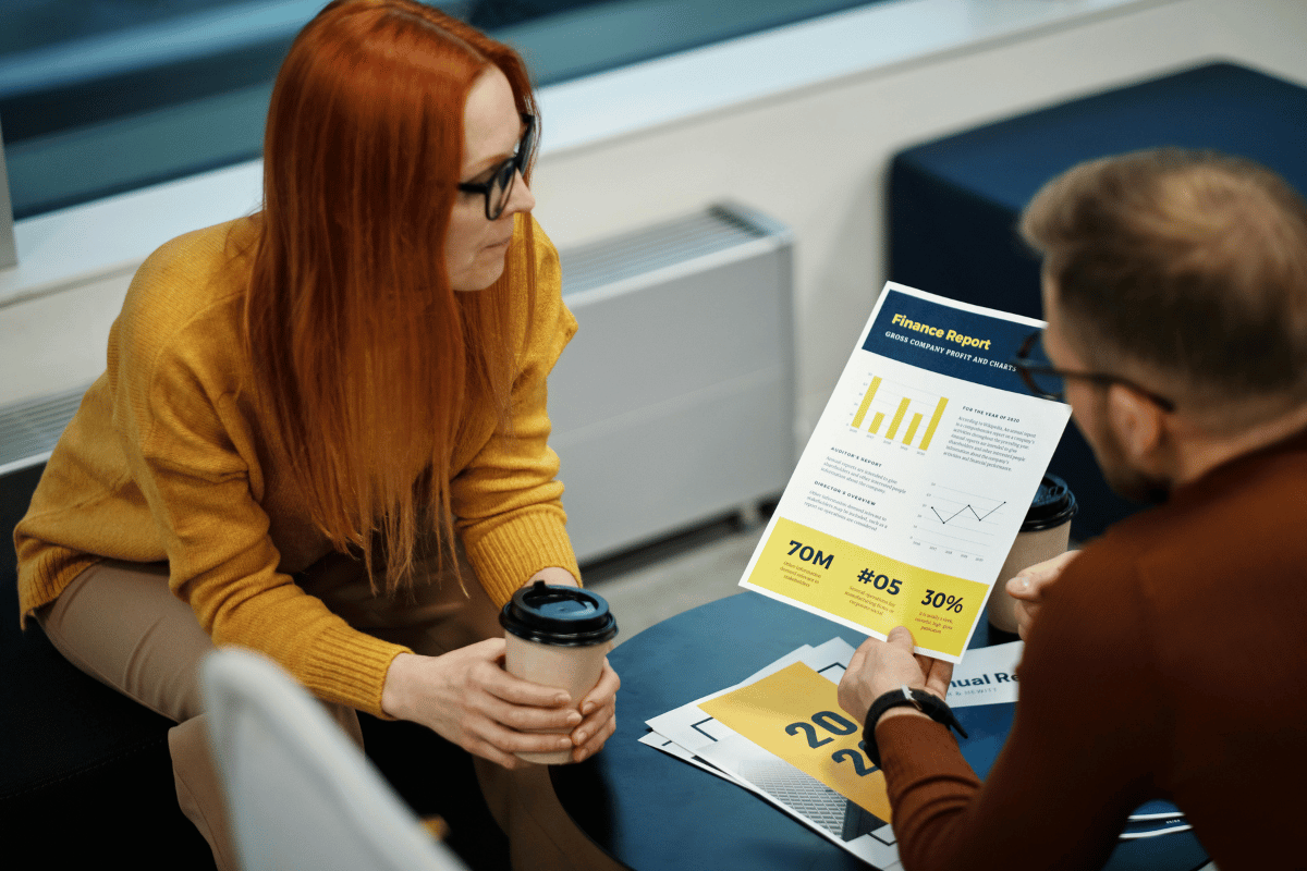 Red-haired woman in glasses talking to a man holding a finance report.