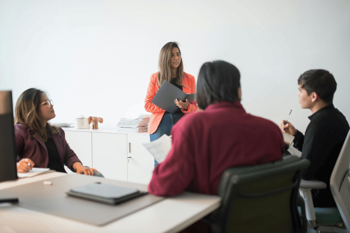 Woman in an orange blazer leading a meeting with three colleagues.