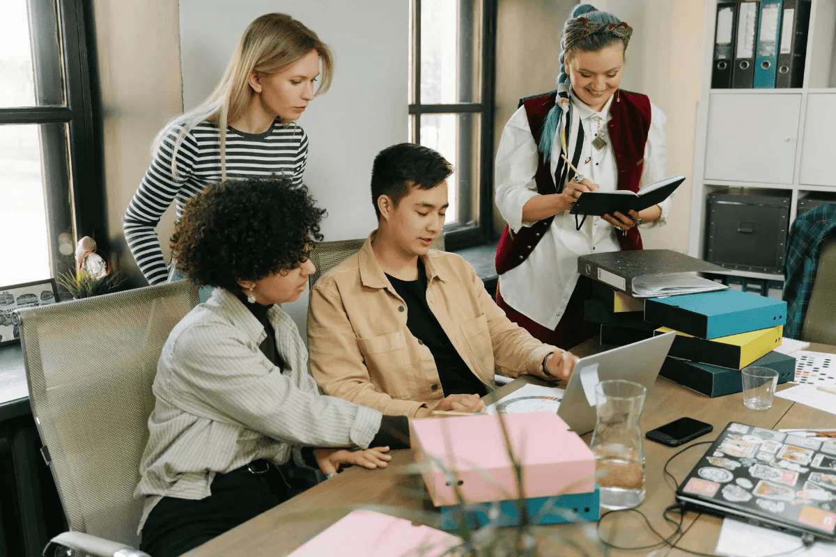 A group of four young professionals collaborating around a table, with one person taking notes in a notebook.