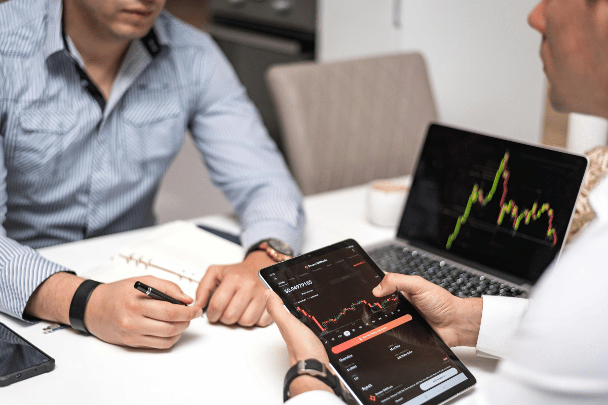 Two professionals reviewing financial data on a tablet and laptop with stock market graphs visible on the screens.