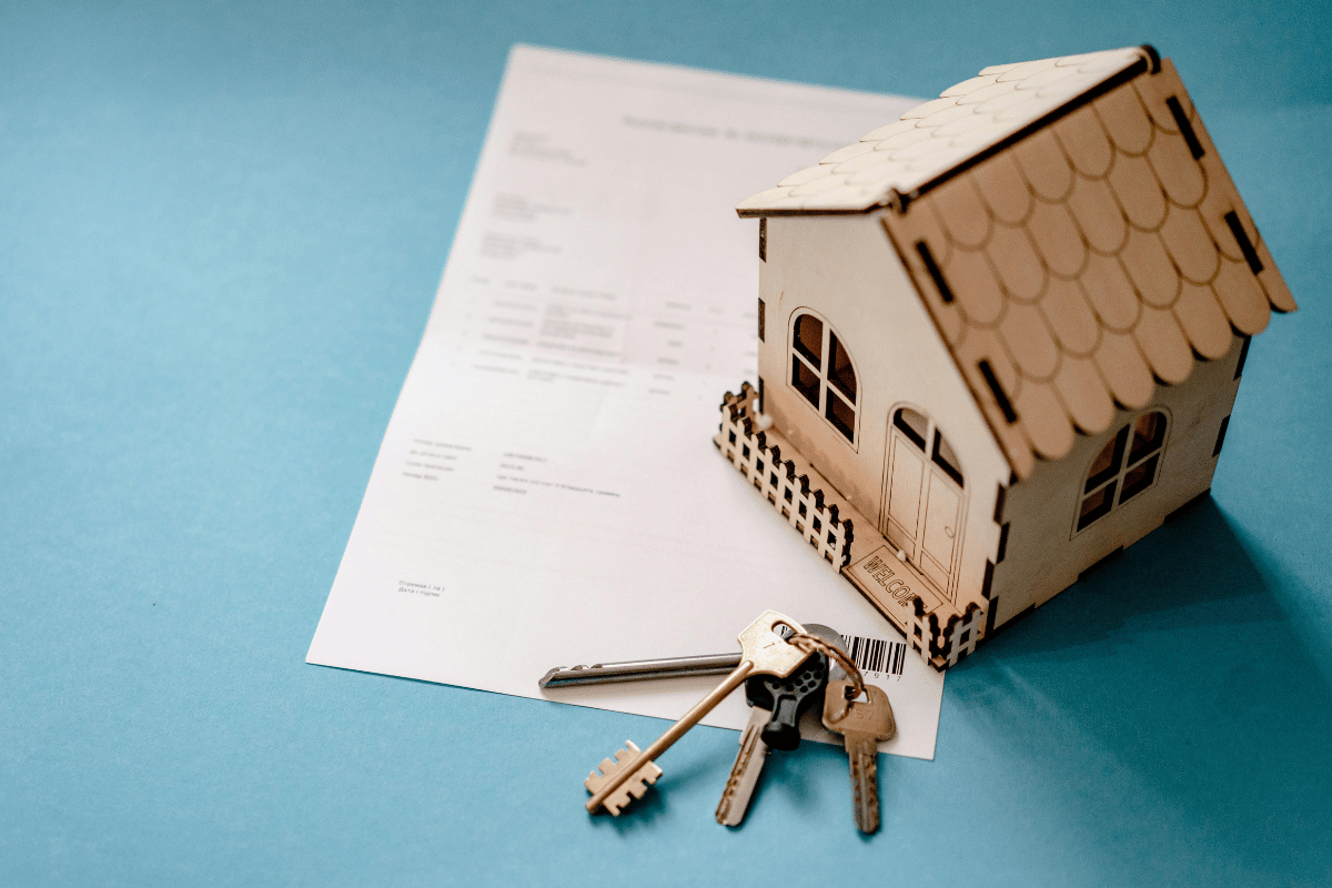 Wooden model house next to property sale agreement with keys on a blue background.