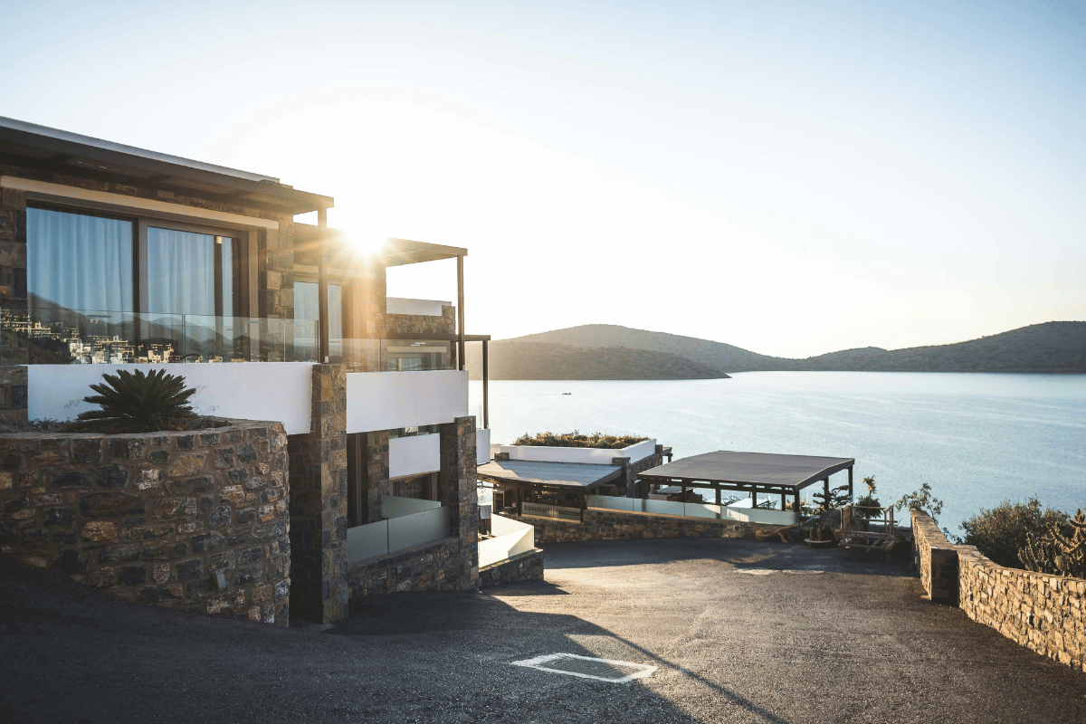 Modern beachfront villa with glass and stone architecture overlooking a tranquil bay at sunset.