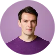 Portrait of a man with short brown hair wearing a purple shirt against a light purple background.