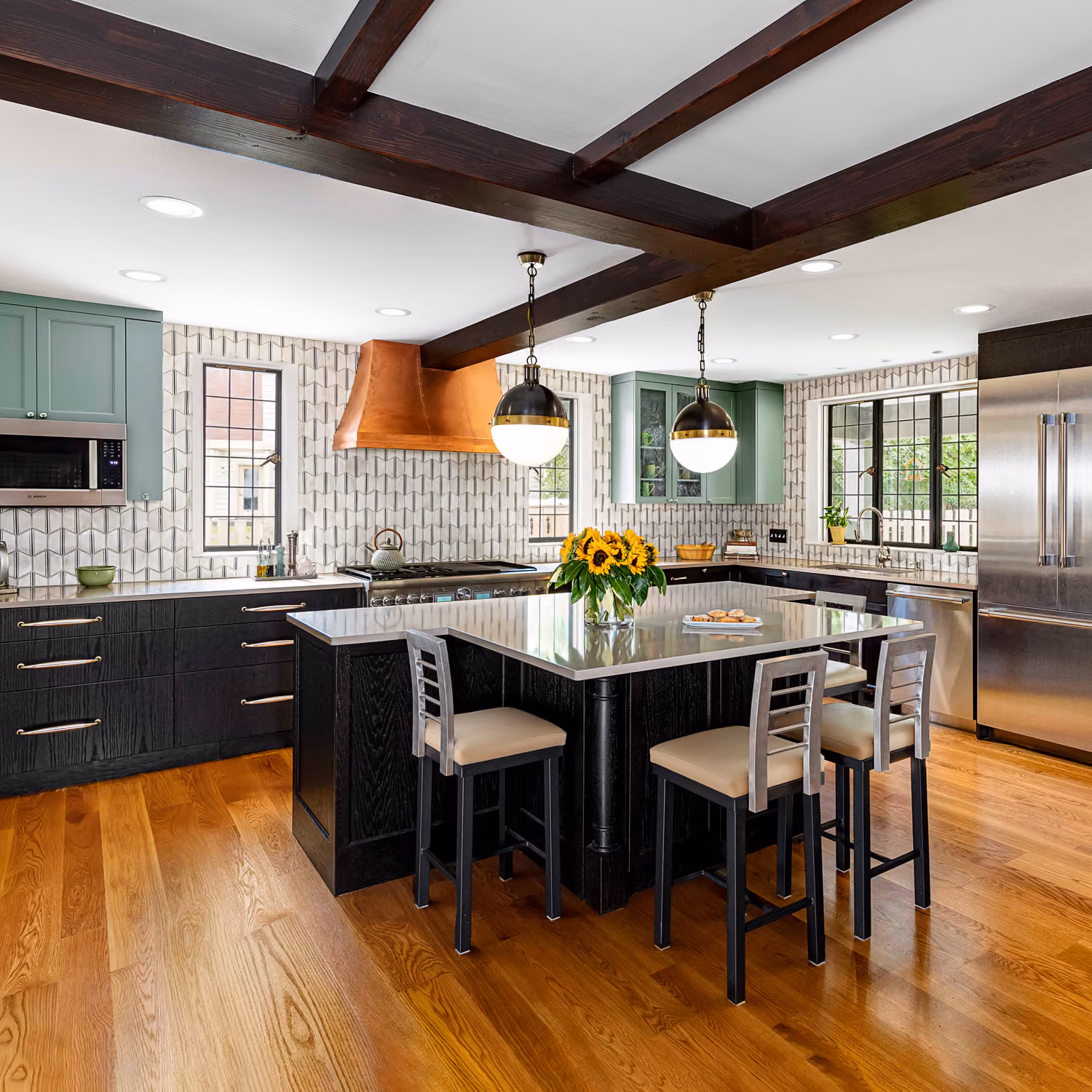 Modern kitchen with wooden floor, dark island, green cabinets, copper hood, and sunflower centerpiece.