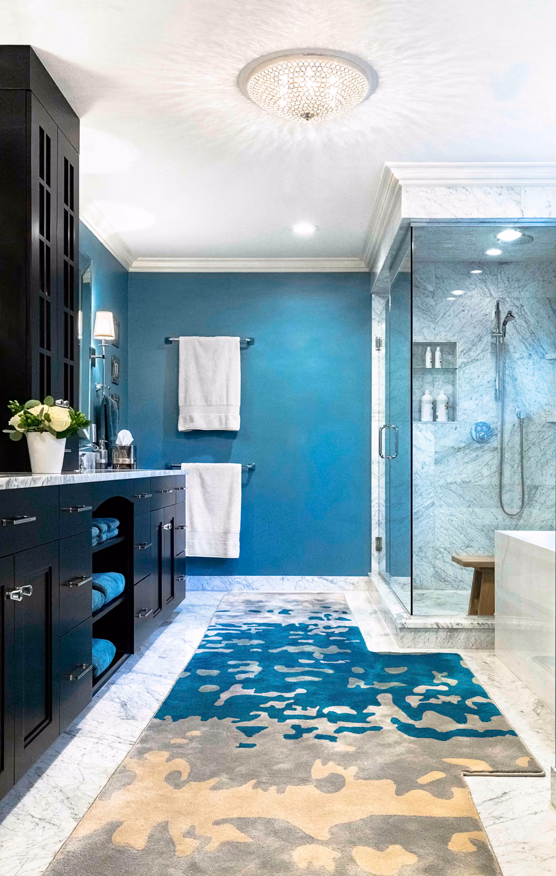 Modern bathroom with blue walls, marble floor, glass shower, dark wood cabinets, and patterned rug.