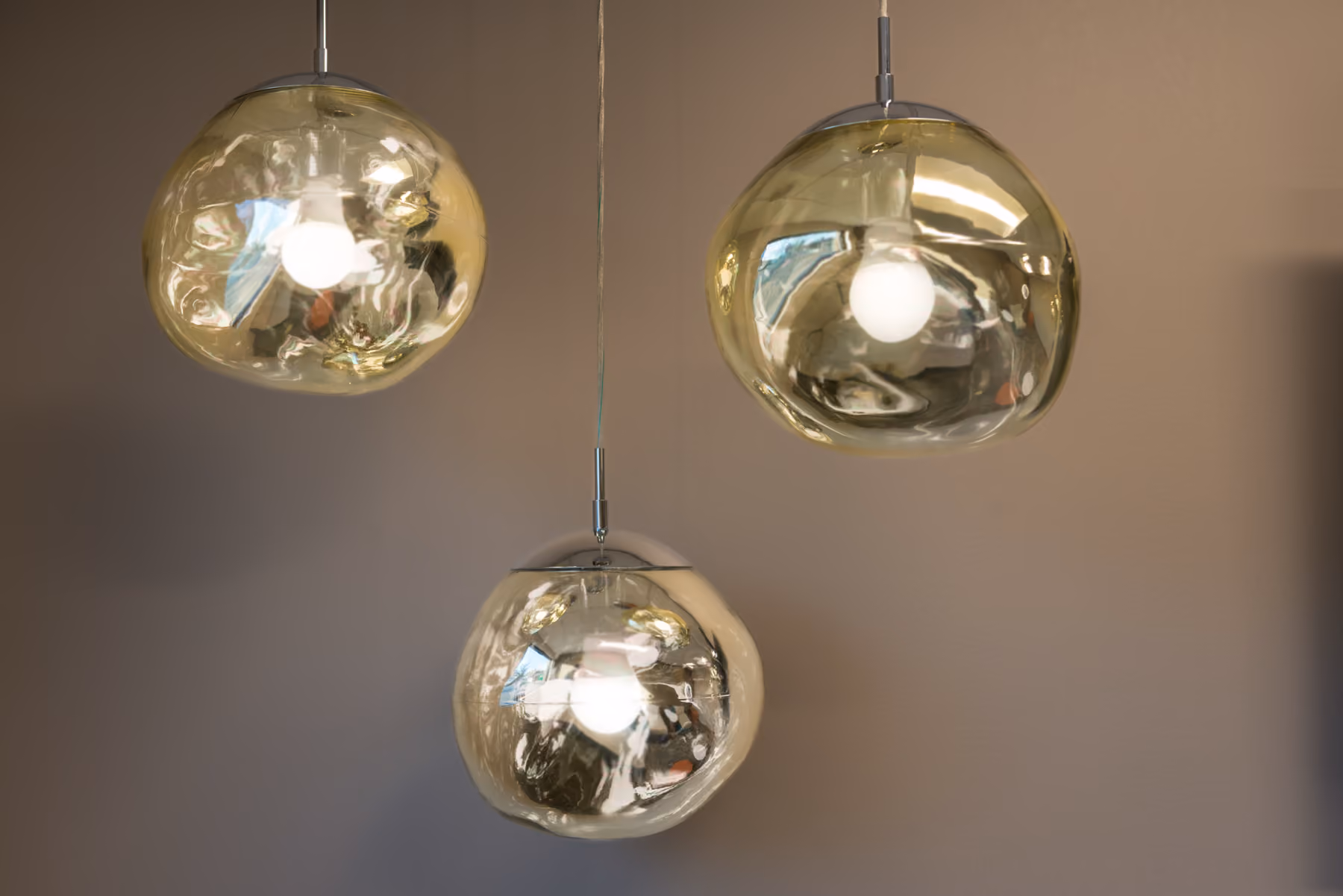 Three modern reflective gold pendant lights hanging against a plain beige wall.