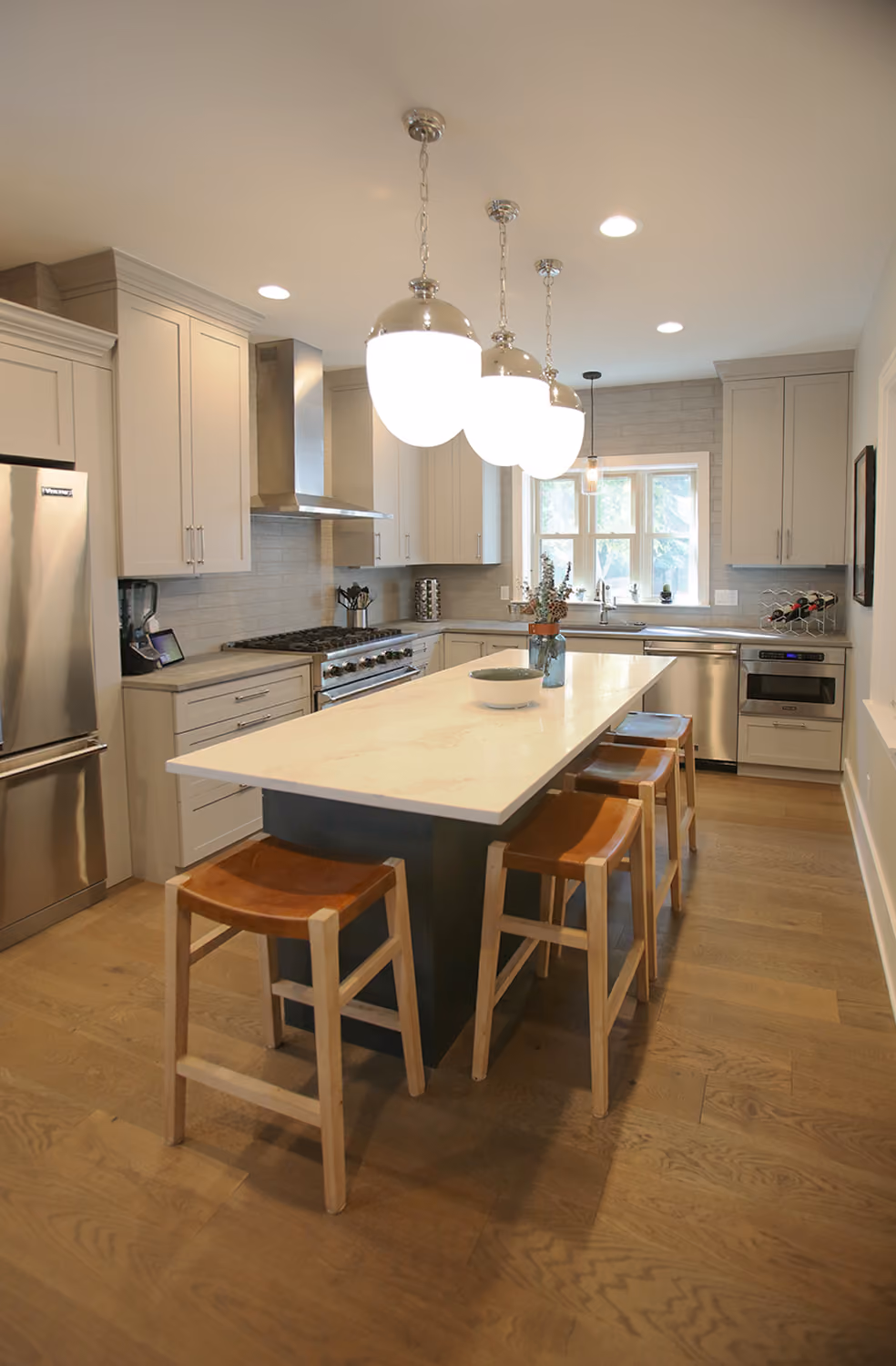 Modern kitchen with white island, wooden stools, stainless steel appliances, pendant lights, and wooden floor.