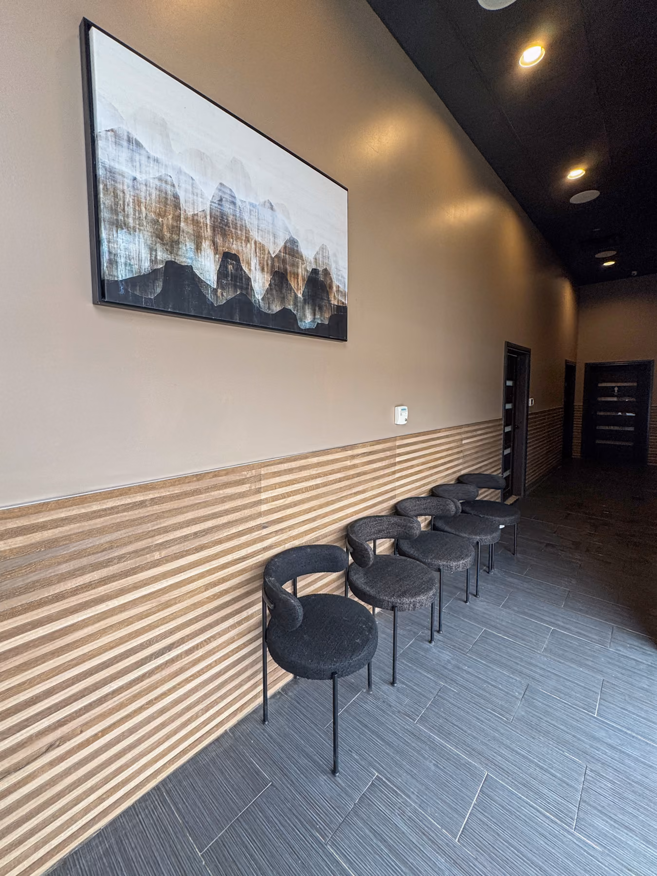 Modern waiting area with six black chairs against striped wooden wall and abstract mountain painting.
