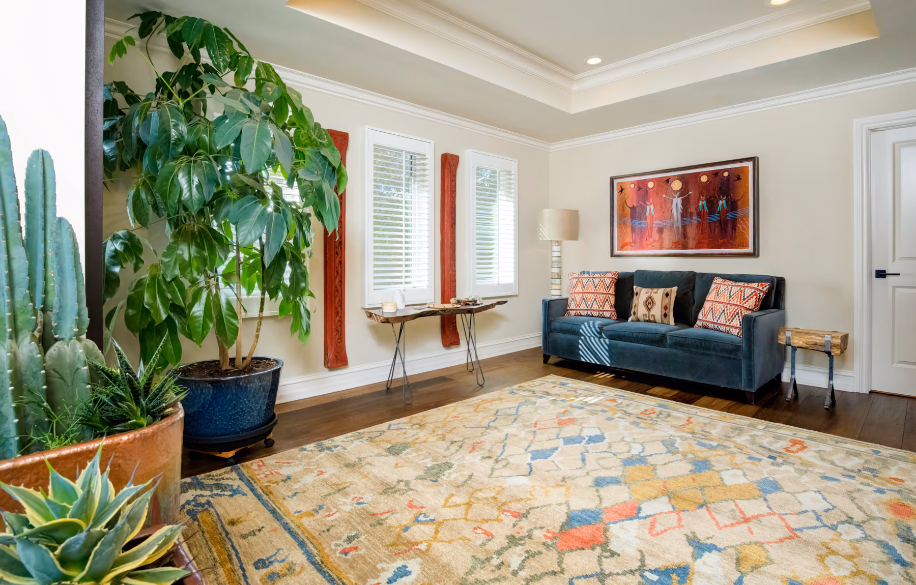 Living room with a blue couch, colorful cushions, large patterned rug, green plants, and wall art.