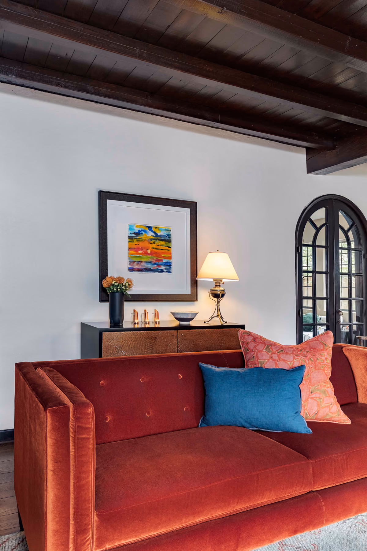 Red velvet sofa with blue and patterned pillows in a living room with lamp and colorful wall art.