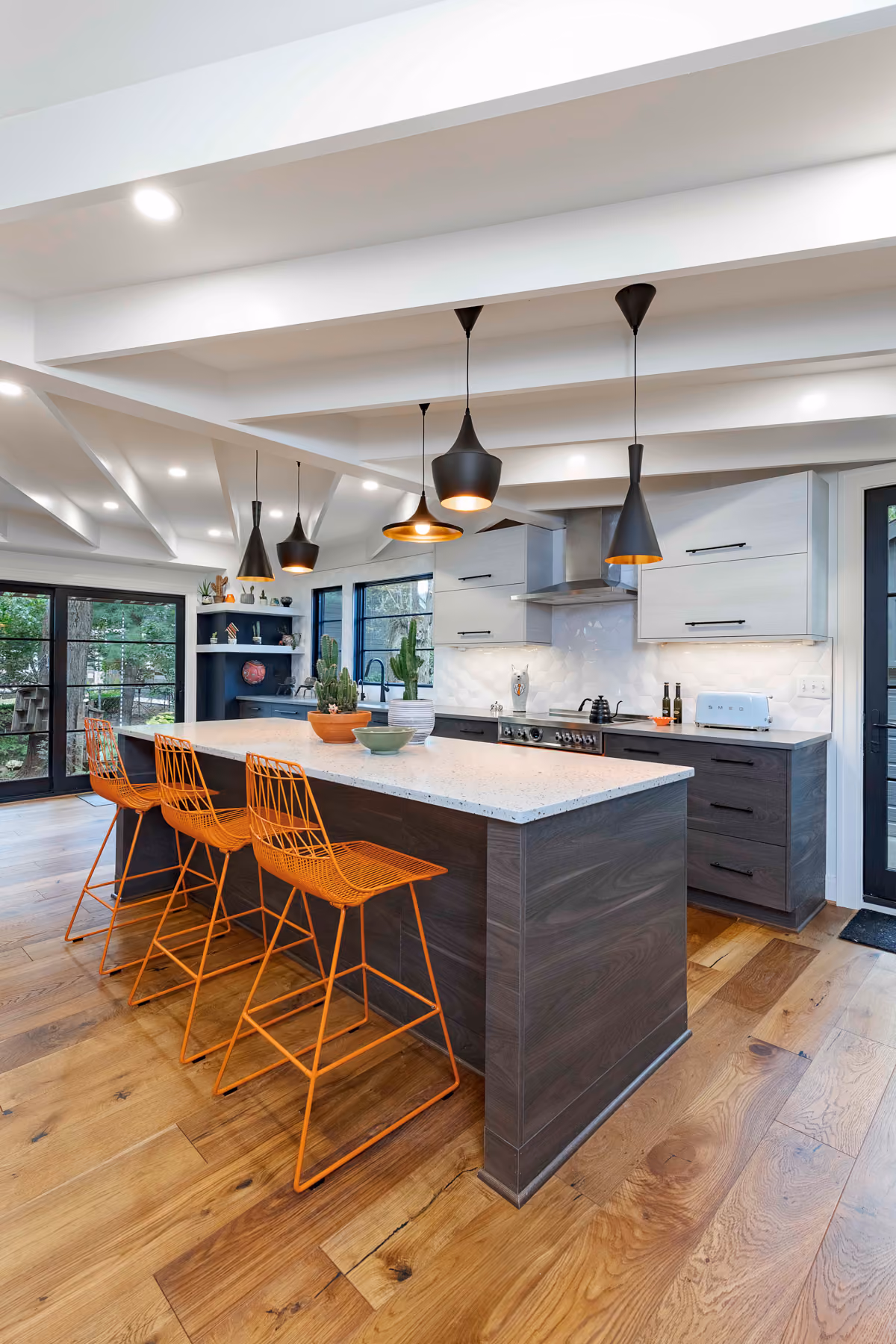 Modern kitchen with wooden island, orange wireframe bar stools, hanging black pendant lights, and hardwood floors.