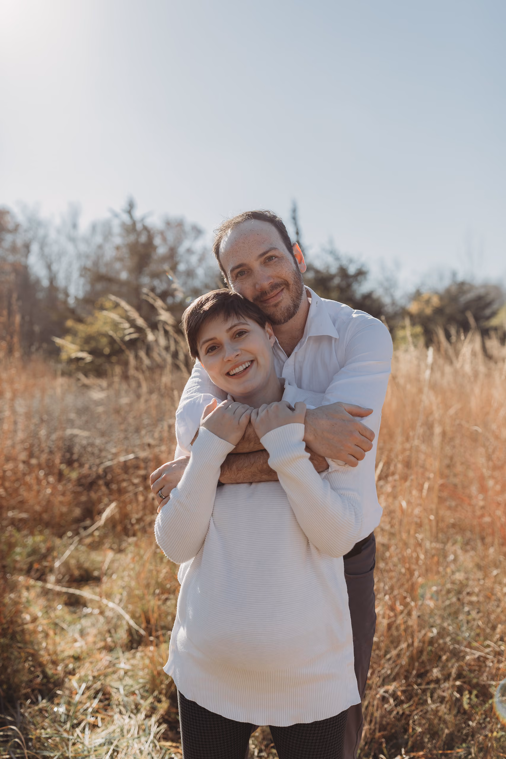 Man hugging smiling pregnant woman from behind in a sunlit field with dry grass and trees.