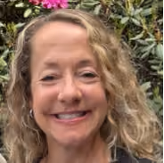 Smiling woman with curly blonde hair outdoors with green leaves and purple flowers in background.