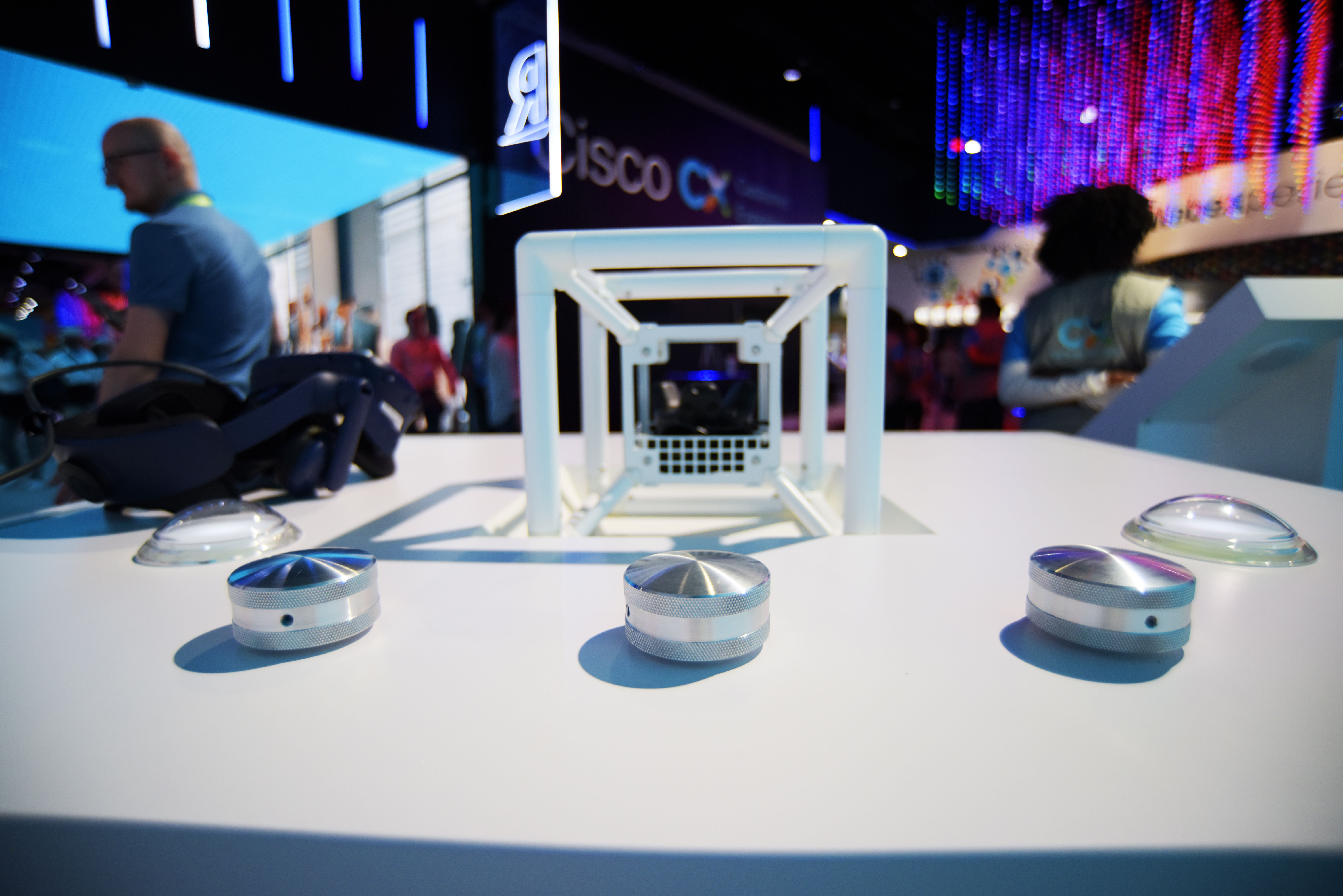 White exhibition table with metal knobs and cube frame at a tech event with people in background.