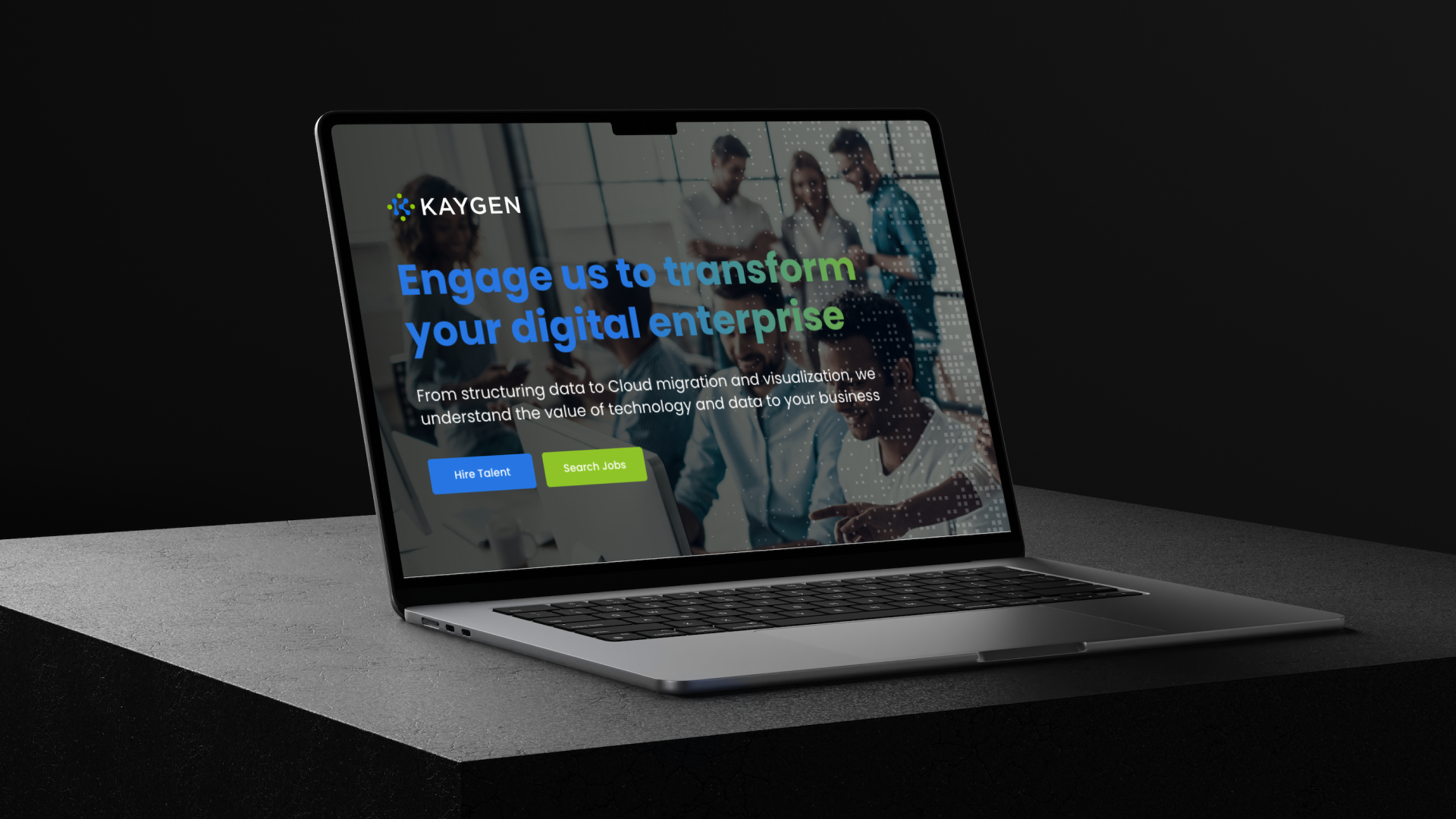 Open laptop on dark textured surface displaying a webpage with KAYGEN logo and text: 'Engage us to transform your digital enterprise.' Below is smaller text about structuring data, cloud migration, and visualization, plus blue and green buttons labeled 'Hire Talent' and 'Search Jobs.' Background shows a blurred group of five people working together.