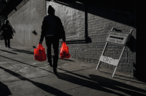A person walks down a sunlit sidewalk carrying red shopping bags