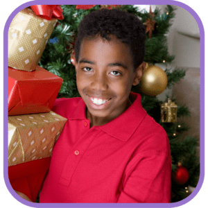 A smiling boy holds wrapped gifts in front of a decorated holiday tree