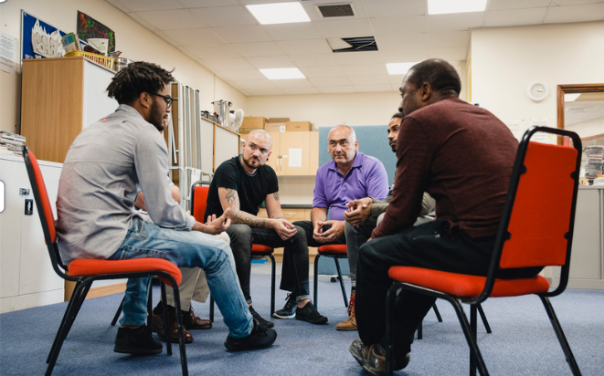 A group of men sit in a circle of chairs talking during a support group