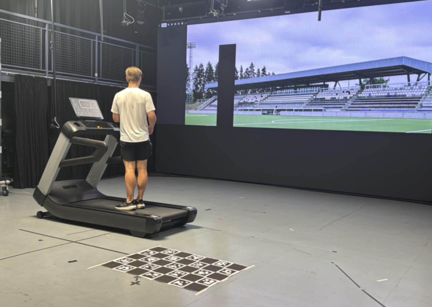 Athlete on a treadmill in the motion-capture lab, facing a screen showing a football stadium, with a calibration board on the floor