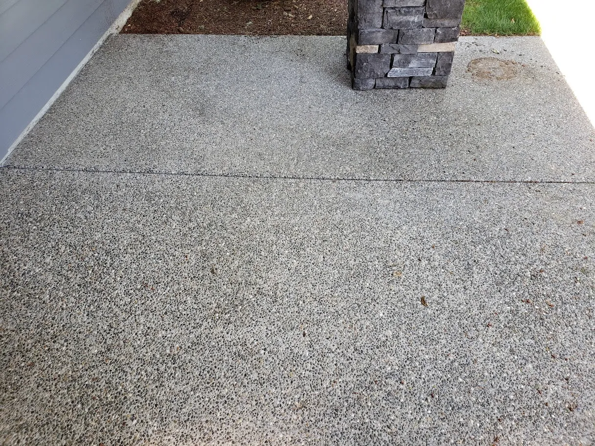Exposed aggregate concrete walkway before restoration, dull grey with a rust-colored stain near the stone column