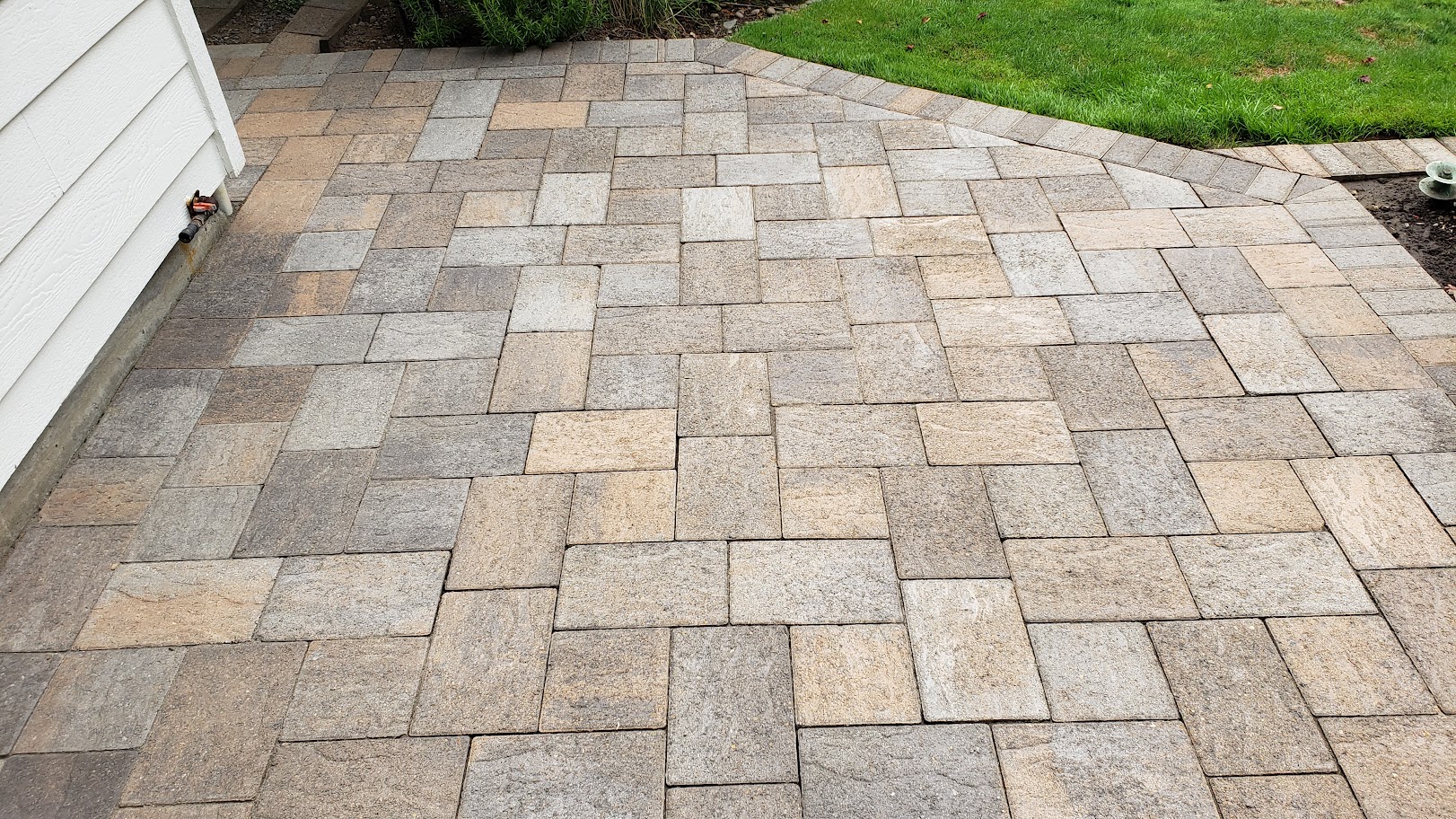 A restored paver patio in tan and grey stone beside a house wall, joints clean and the lawn edge sharp