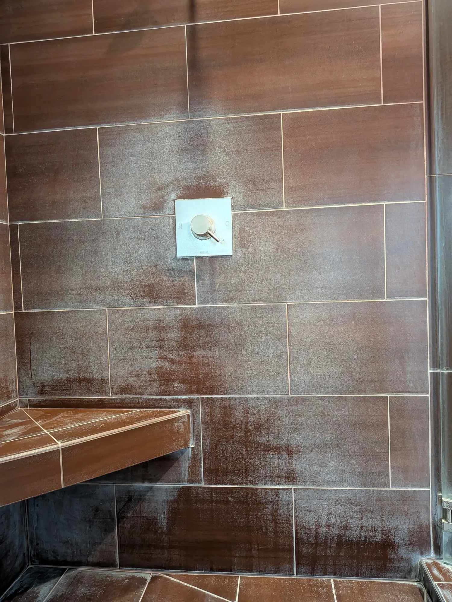A brown tiled shower wall before restoration, the tile and bench coated in a heavy white hard water film