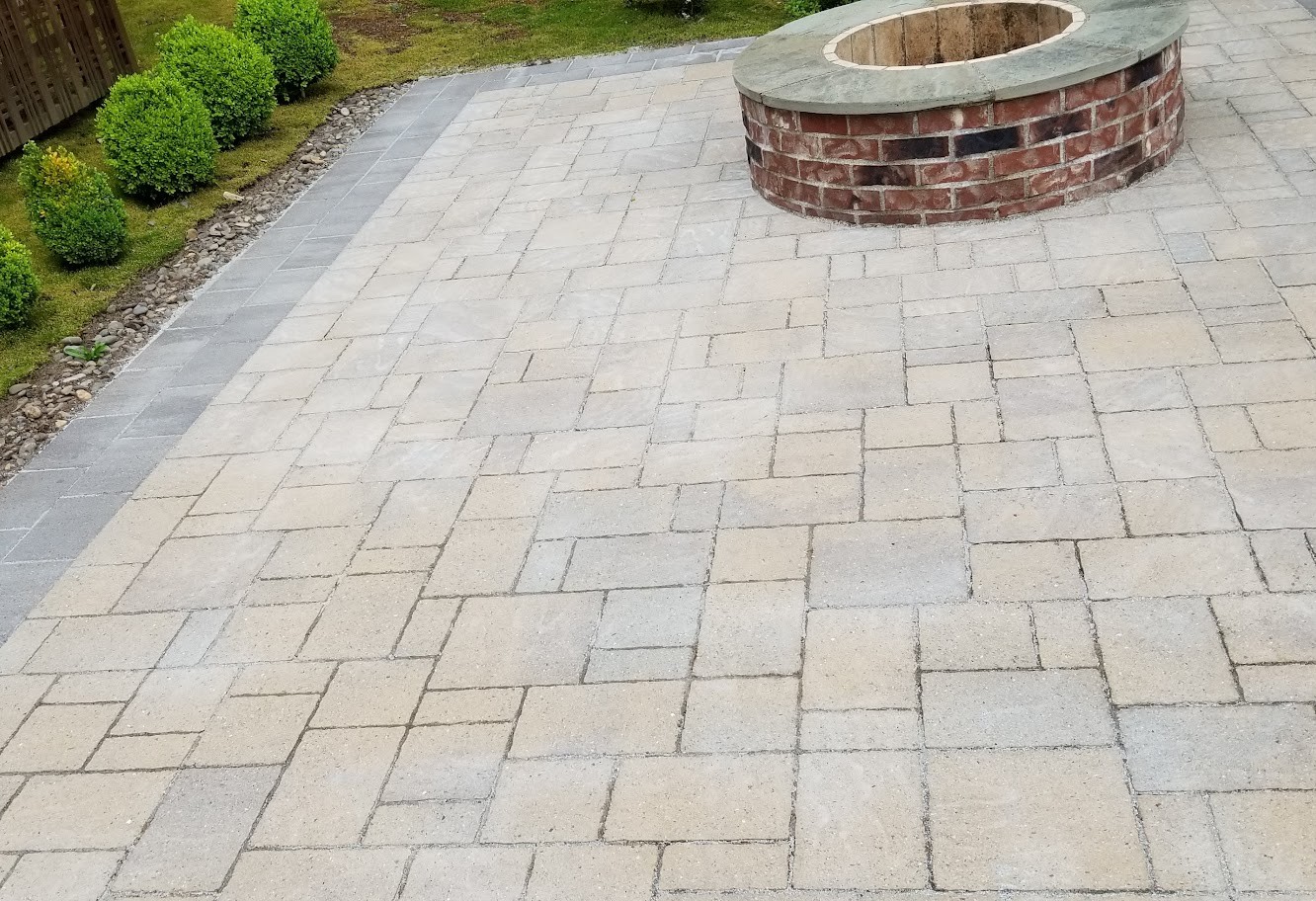 A restored paver patio in a back garden, clean and even in tone, curving around a brick fire pit with a clipped hedge and fence behind