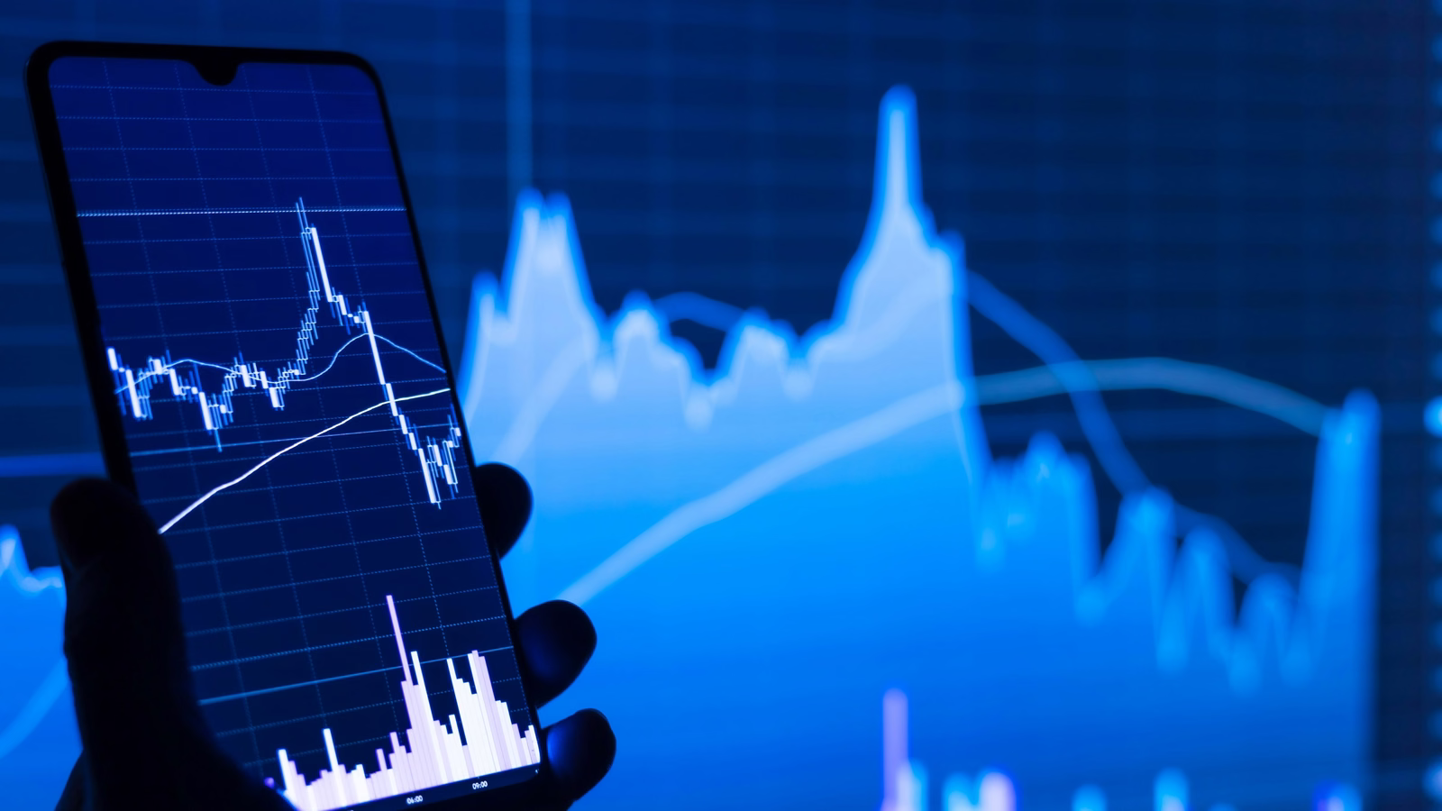 Hand holding a phone with a rising chart in front of a larger screen of charts