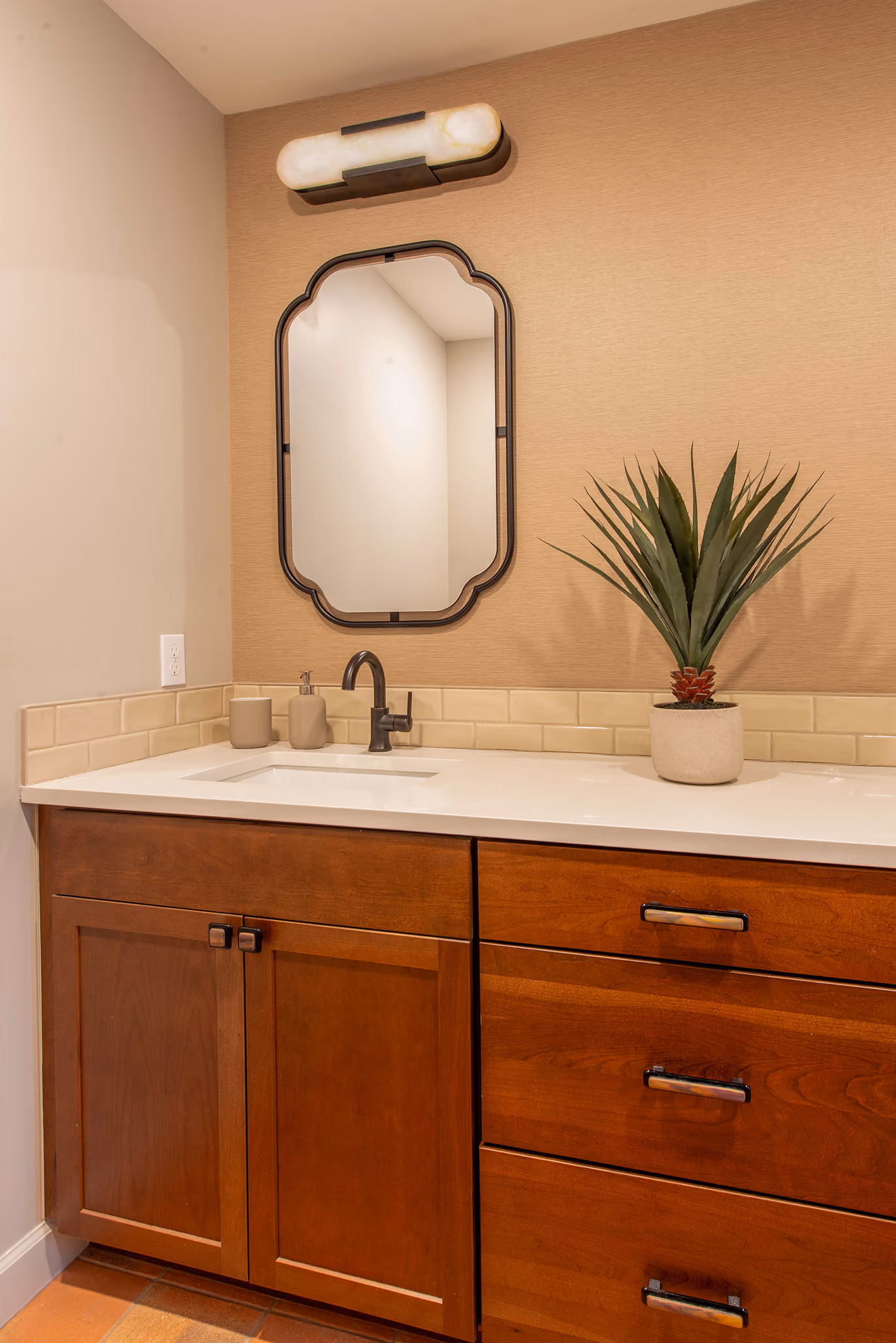 Powder room vanity with warm wood cabinetry, an arched black-framed mirror, and patterned wallpaper