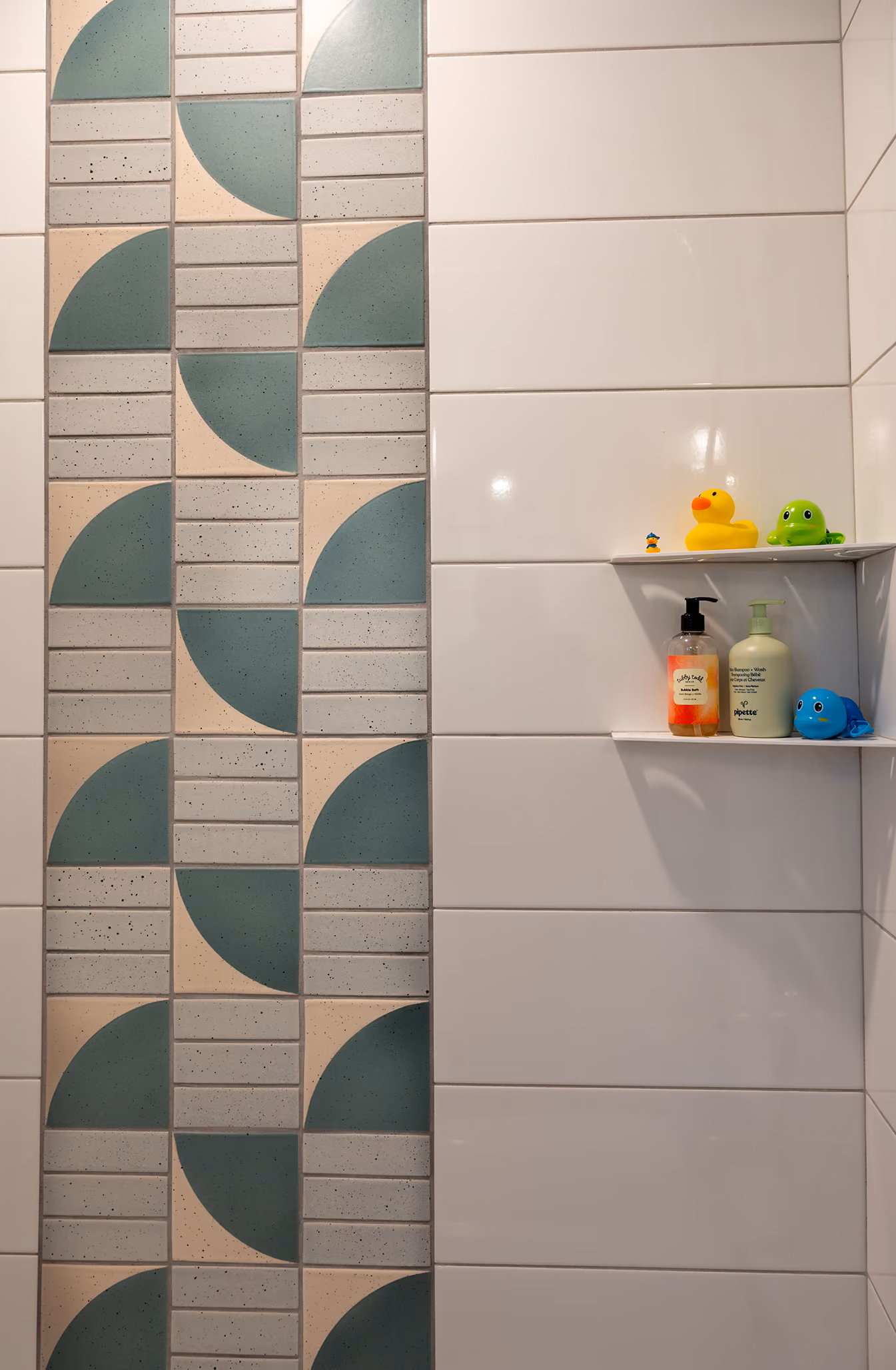 Kids' bathroom shower with geometric fan-pattern tile and rubber duck toys on a built-in niche shelf
